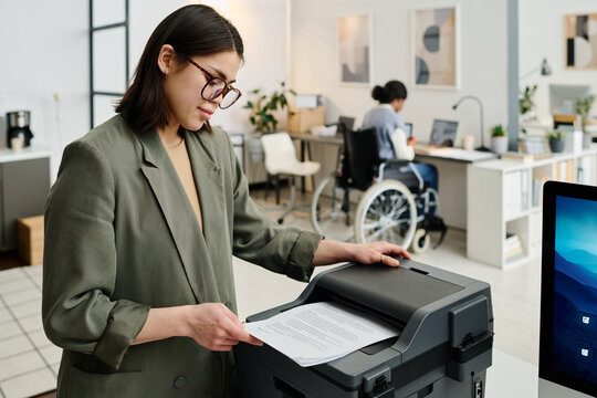 Young female manager wearing stylish eyeglasses printing documents, her colleague with disability working in background