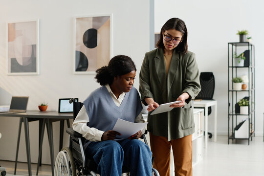 Medium long shot of young Caucasian woman showing data on digital tablet to her African American coworker with disability - Powered by Adobe