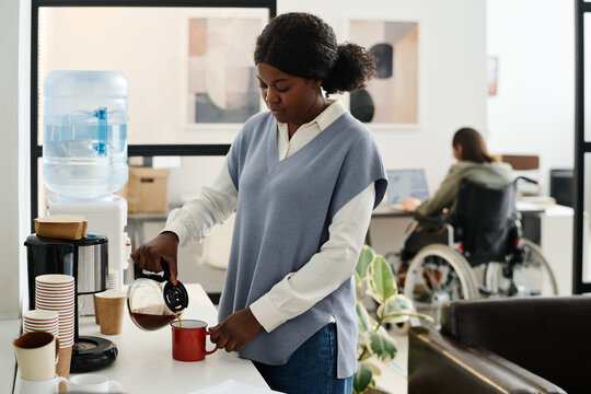 Selective focus shot of young African American woman pouring coffee into mug during break at work in modern office