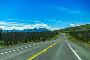 Scenic Highway in Alaska to the Alaska Range mountains on horizon at the end of long highway with green trees on side of highway. 