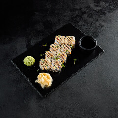 Sushi roll with vegetables and fried salmon inside, sesame seeds outside, and spicy sauce, presented on a black slate with soy sauce, wasabi, and ginger