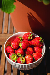 Srawberries in a white ceramic bowl on the wooden terrace table. Natural light. Outdoors. 