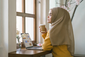 Fototapeta na wymiar A thoughtful Muslim woman holding a coffee cup gazes out a window, taking a break from work at her laptop.