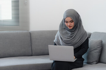 Muslim woman in a hijab concentrating on her work on a laptop while sitting on a sofa in a home.