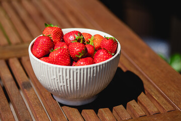 Srawberries in a white ceramic bowl on the wooden terrace table. Natural light. Outdoors. 