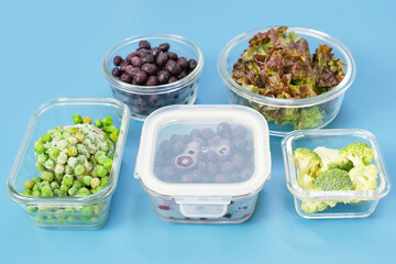 Green vegetables and berries in glass lunch boxes on blue background close-up, side view.
