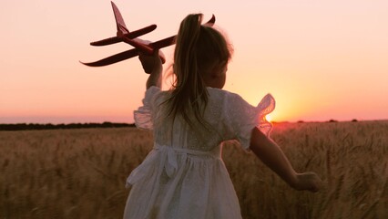 Happy little girl kid in white dress running with plane toy on wheat field at sunset sunrise...