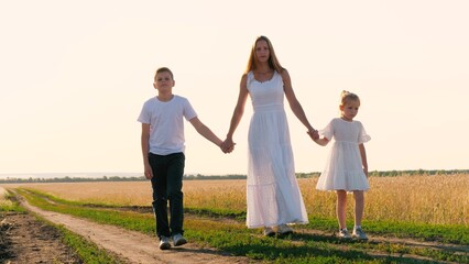 Elegant young mother walking with son and daughter on dirt road sunny summer field holding hands enjoy motherhood. Happy family going together outdoor countryside sunset sunrise nature landscape