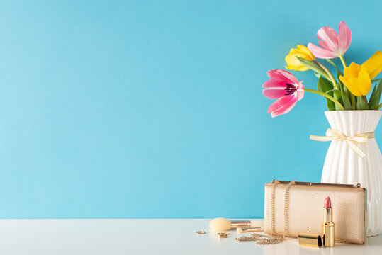 Mother's Day and 8 March theme. Table side view featuring women's essentials like gold jewelry, makeup brushes, lipstick, designer bag, and tulips in a vase against a soft blue backdrop for text