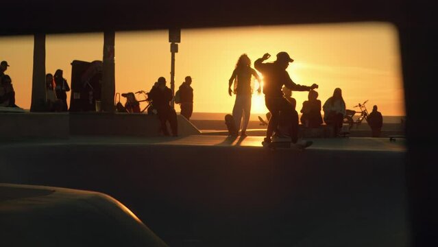 Silhouettes of skateboarders hanging out and riding skateboards in front of sunset sun at Venice Beach skate park in Los Angeles, California. Slow motion shot.