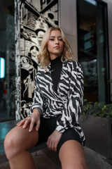 Fototapeta na wymiar Fashionable urban girl model in a stylish zebra print dress with sports shorts sits in the city near a building