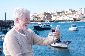 Video call concept. Senior smiling bearded man sitting at sea harbour talking on mobile phone using...
