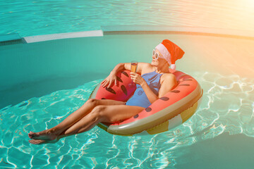 A happy woman in a blue bikini, a red and white Santa hat and sunglasses poses in the pool in an inflatable circle with a watermelon pattern, holding a glass of champagne in her hands. Christmas