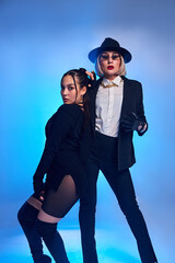 female modern dancers in elegant suit and dress pose in dance move. Two attractive women dance together, posing at camera, sexy and gorgeous. isolated in studio, with blue backlit in background