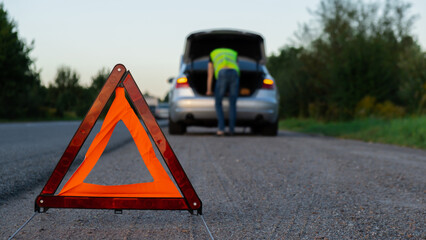 Broken silver luxury car emergency accident. Man driver installing red triangle stop sign on road. Sport automobile turned on blinkers technical problems on the road. Safety procedure vehicle broken 