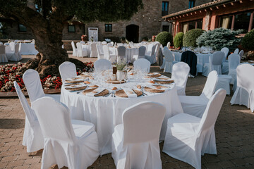 
Banquete de bodas en el exterior de la masía. Mesas decoradas con manteles blancos, sillas blancas, cristalería transparente. Entorno natural, olivo, piedra y adoquines. Día de verano.
