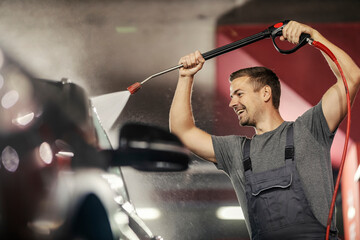 A cheerful worker is washing a car with high pressure jet gun at station.