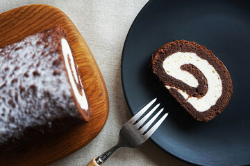 Chocolate sponge roll with vanilla cream on a wooden board next to a cut piece of rolls on a plate on a light background