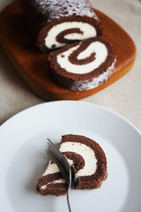 Chocolate sponge roll with vanilla cream on a wooden board next to a cut piece of rolls on a plate on a light background