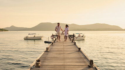 couple walking on a wooden pier in the ocean at sunset in Thailand.Caucasian men and Asian woman diverse couple waking at a jetty in the ocean, man and women watching sunset together