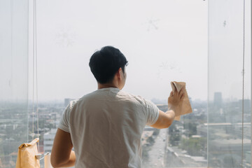 Backside view of asian Thai man wiping window glass in room apartment with city view, keep glass...