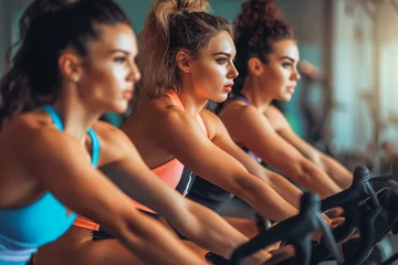 Plexiglas foto achterwand Group of three sporty women in sportswear riding stationary bikes on cycling class at gym. © VisualProduction