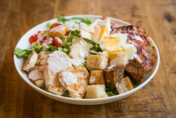 Close up of chicken Caesar salad with bacon, croutons, tomatoes, parmesan cheese, boiled eggs and dressing in a white bowl on a wooden table