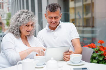 Attractive elderly family couple sitting in sidewalk cafe using tablet computer.