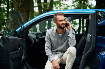 Talking by phone. Beautiful young man is sitting in the blue car outdoors