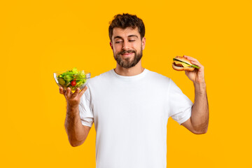 Smiling Caucasian Man Holding Cheeseburger And Healthy Vegetable Salad, Studio