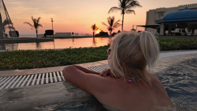Scenic view of a woman savoring a sunset beside the pool. Serene interlude in the twilight, as a girl unwinds in an infinity pool, captivated by the stunning horizon on a tropical beach.