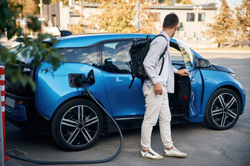 With backpack. Man with blue electric car on the charge station