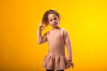 Smiling child girl showing ok gesture over yellow background. Positive emotions concept