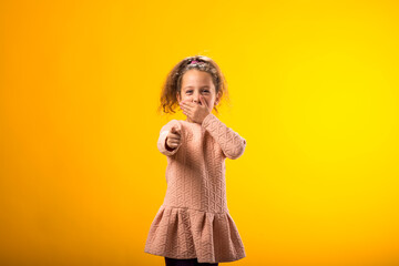 Kid girl mocking and teasing at someone showing finger at camera and closing her mouth over yellow...