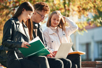 Learning, sitting and using laptop. Three young students are outside the university outdoors