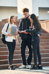 Holding notepads in hands. Three young students are outside the university outdoors