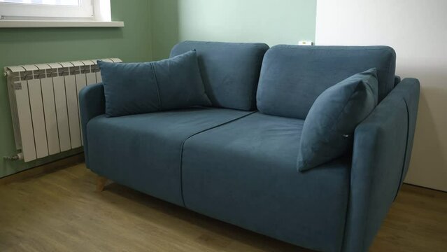 Blue sofa with soft cushions and velvet upholstery stands by window in room corner. Cosy couch for visitors and employees rest in office