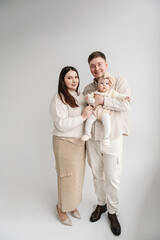 Family. Father, mother hugging daughter. Happy family hugs cute baby on a holiday. Dad, mom holds in hands 6 month old little kid. Daddy, mommy embracing girl with bow in hair isolated on white wall.