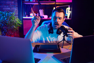 Young man, broadcaster in casual T-shirt expressing ideas into microphone in neon-lit room at home-studio. Concept of youth people with social media and smart working, lifestyle, online streaming.