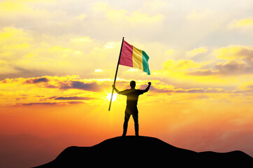 Guinea flag being waved by a man celebrating success at the top of a mountain against sunset or sunrise. Guinea flag for Independence Day.