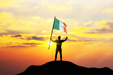 Ireland flag being waved by a man celebrating success at the top of a mountain against sunset or sunrise. Ireland flag for Independence Day.