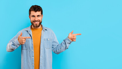 Smiling bearded millennial guy points fingers to side, blue backdrop