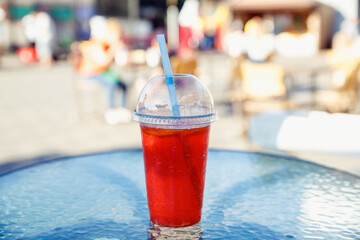 Summer strawberry red lemonade in plastic cup with lid and cocktail straw on glass table of sidewalk cafe.