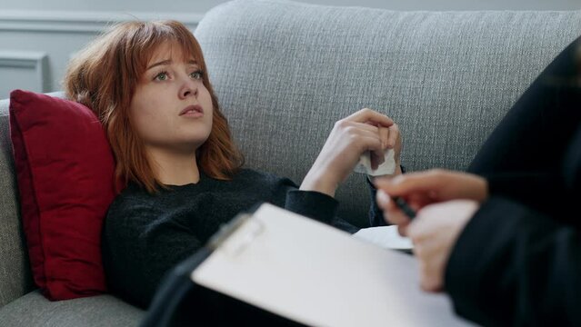 Frustrated young red hair girl laying down the couch and telling sad news her psychotherapist. Nervous lady giving feedback for professional psychological consultant indoors