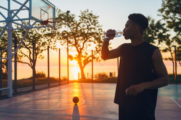 cool black man doing sports in morning, drinking water on basketball court on sunrise