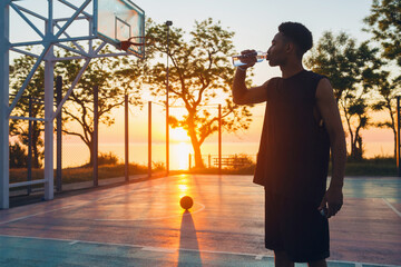 black man doing sports in morning, drinking water on basketball court on sunrise