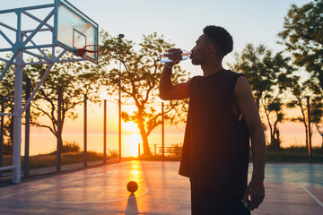 black man doing sports in morning, drinking water on basketball court on sunrise