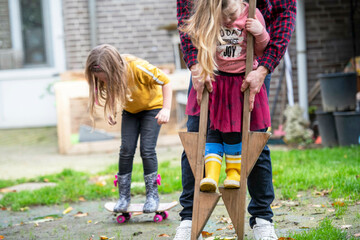 Father helping his daughter balance on stilts in their garden having fun. Her sister in the background trying to skateboard