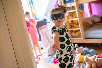 Young girl in costume playing superhero dressing up with her sister in the background