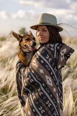 Portrait of a woman in a poncho and a hat with her beloved dog. Summer landscape, feather grass field, sky with clouds.